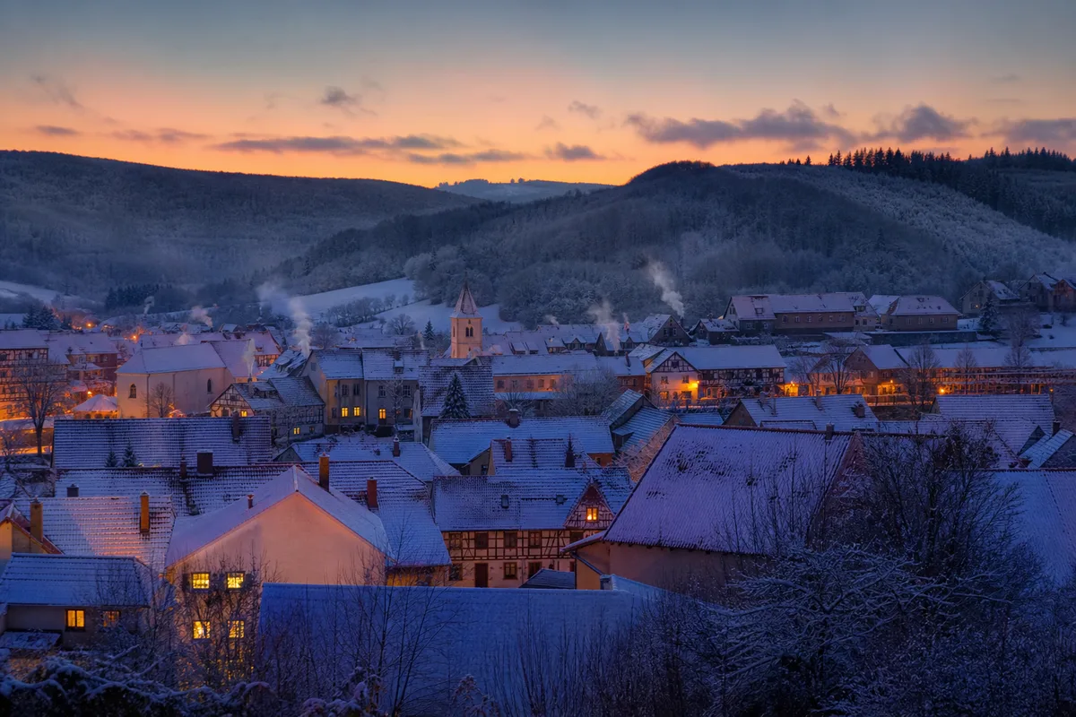 Village lorrain sous la neige, toits de tuiles rouges et cheminées fumantes au crépuscule